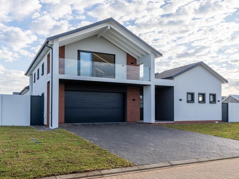 Modern double-story residential home featuring a paved driveway, integrated double garage, and a prominent upper-level balcony with glass balustrades.