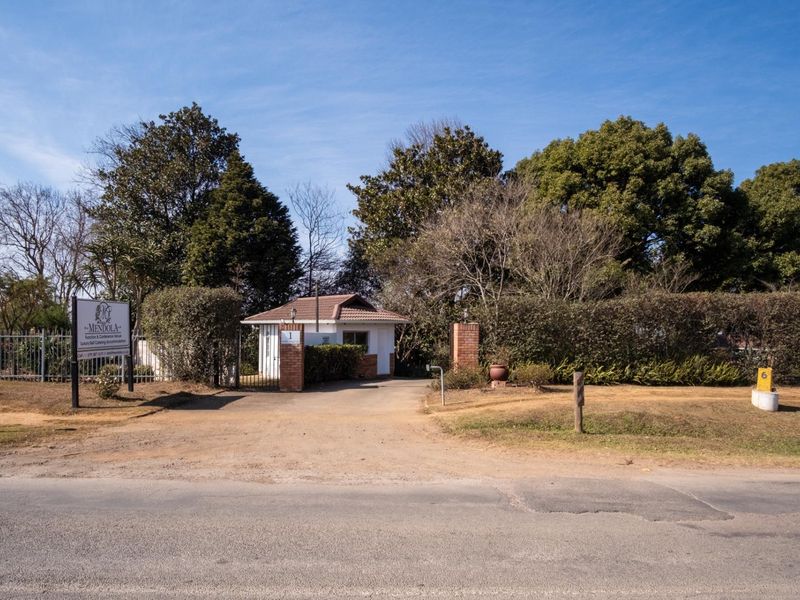 Front entrance view of the property featuring a guard house, brick pillars, security gate, and established garden surroundings.