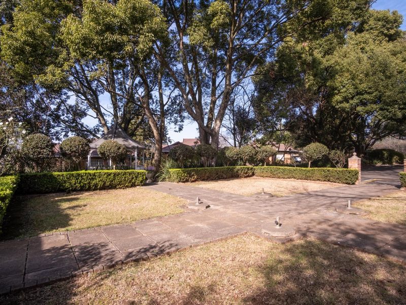 A spacious garden area featuring mature trees, manicured hedging, a paved walkway, and a gazebo structure in a tranquil setting.