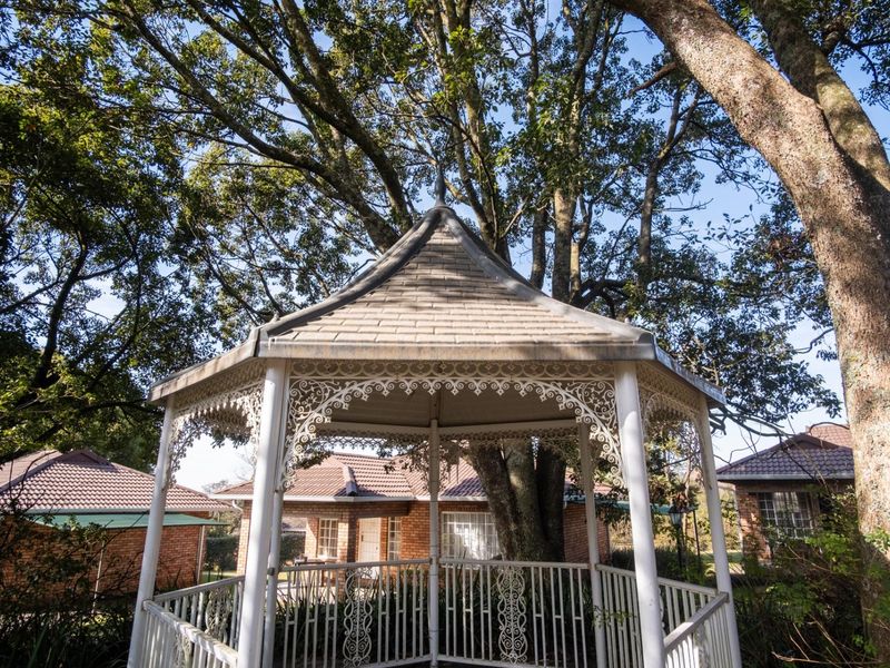A charming white wrought iron gazebo set within a lush, tree-filled garden area, providing a peaceful outdoor space.
