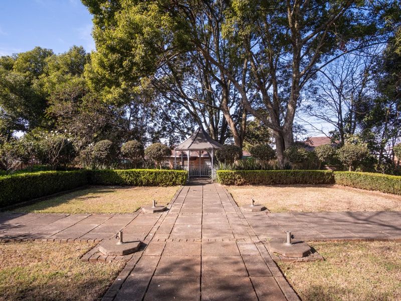 A landscaped garden featuring a central paved walkway leading to a gazebo, surrounded by manicured hedges and mature trees.