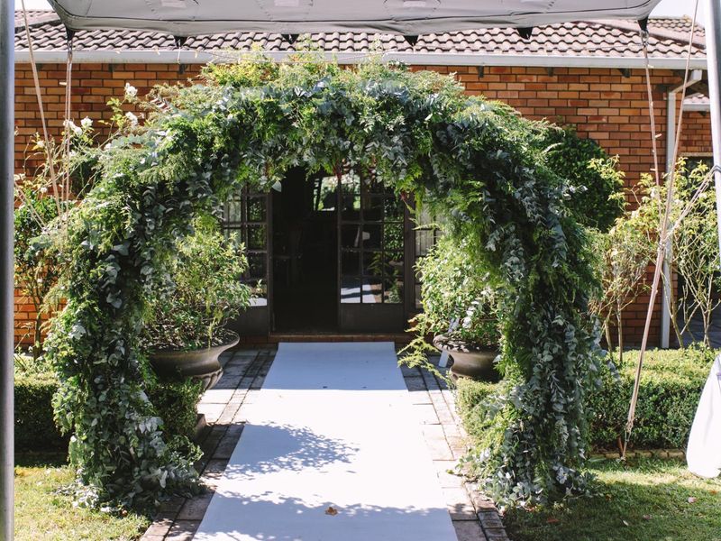 A rear exterior view of a brick building featuring a paved walkway leading to classic french doors, framed by a lush garden archway.