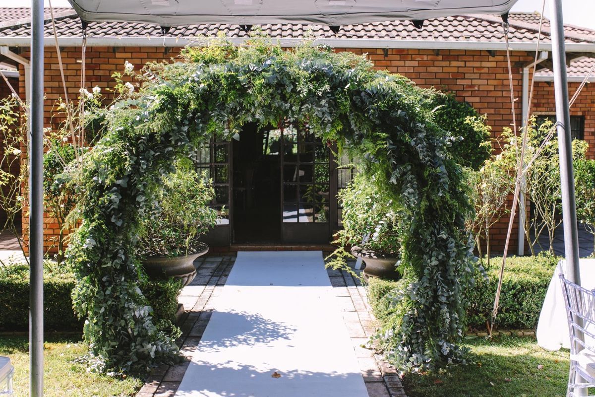 A rear exterior view of a brick building featuring a paved walkway leading to classic french doors, framed by a lush garden archway.