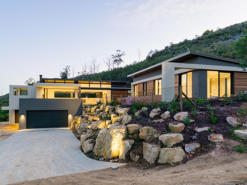 Modern architectural home featuring a paved driveway, integrated double garage, and tiered rock garden landscaping set against a hillside backdrop.