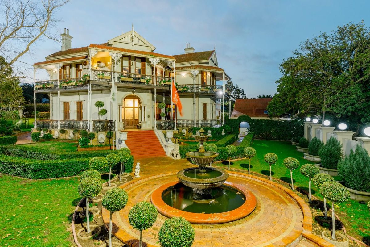 A grand historic residence featuring an expansive landscaped garden, a central ornate fountain, and a classic wrap-around veranda with intricate wrought iron detailing.