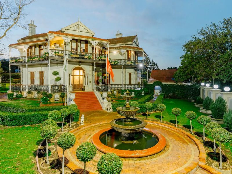 A grand historic residence featuring an expansive landscaped garden, a central ornate fountain, and a classic wrap-around veranda with intricate wrought iron detailing.