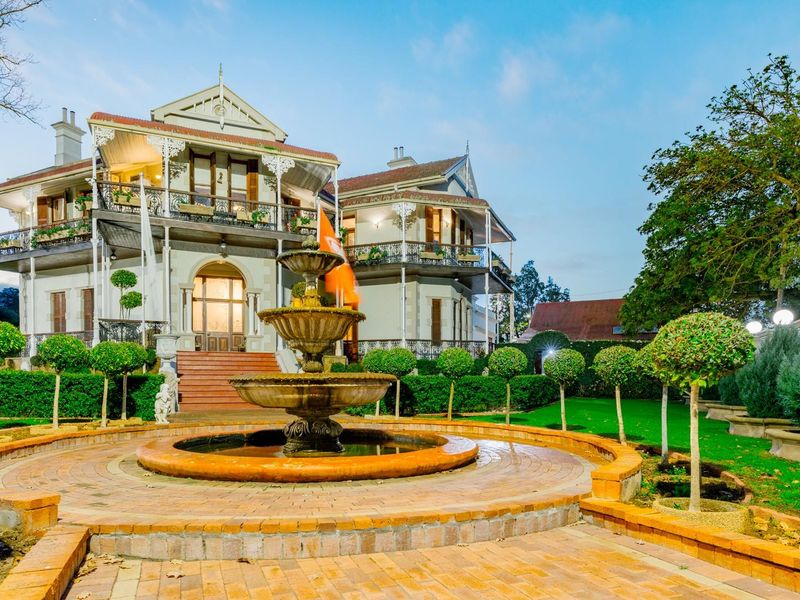 A grand historic residence featuring a tiered stone fountain, manicured gardens, and expansive two-story verandas with intricate wrought iron detailing.