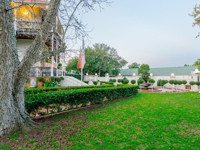 A well-maintained garden featuring a central fountain, manicured hedges, and stone boundary walls, adjacent to a multi-story residence with a wrought iron balcony.