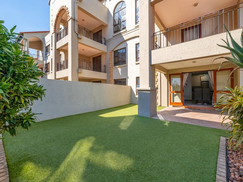A well-maintained rear garden area featuring artificial turf, a paved patio, and access to the apartment's ground floor living space and upper-level balconies.