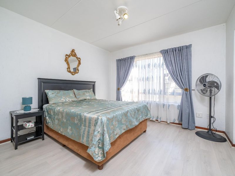 A well-lit bedroom featuring light-toned laminate flooring, neutral walls, and a large window dressed with curtains.