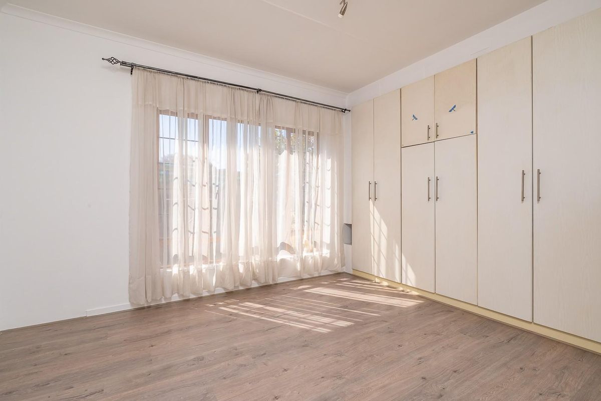 A bright, empty bedroom featuring light-toned built-in cupboards, modern laminate flooring, and large windows with installed curtain rods.