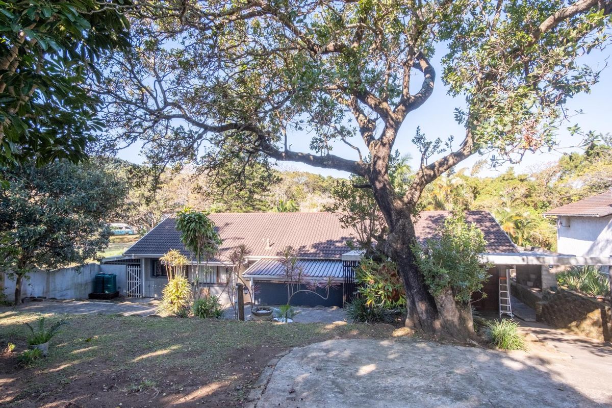 A view of the rear exterior of the house featuring a tiled roof, concrete driveway, and established garden with mature trees.