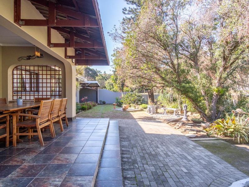A spacious covered patio featuring tiled flooring and exposed timber trusses, overlooking a well-established garden and paved outdoor area.