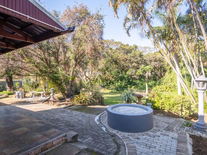 A spacious rear garden featuring a paved patio area, a covered veranda, and a built-in circular fire pit surrounded by mature trees and landscaping.