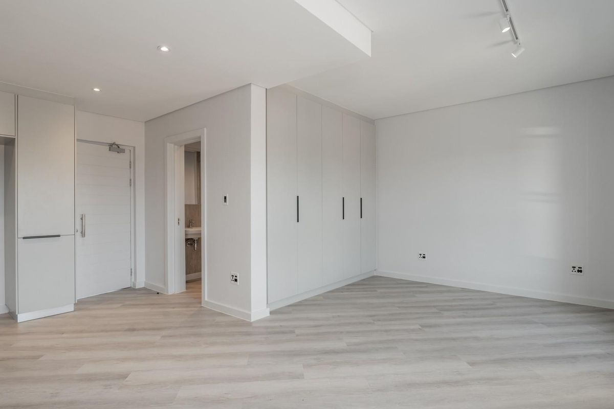 A bright, modern, and empty bedroom featuring neutral laminate flooring, ample built-in cupboard storage, and contemporary recessed and track lighting.