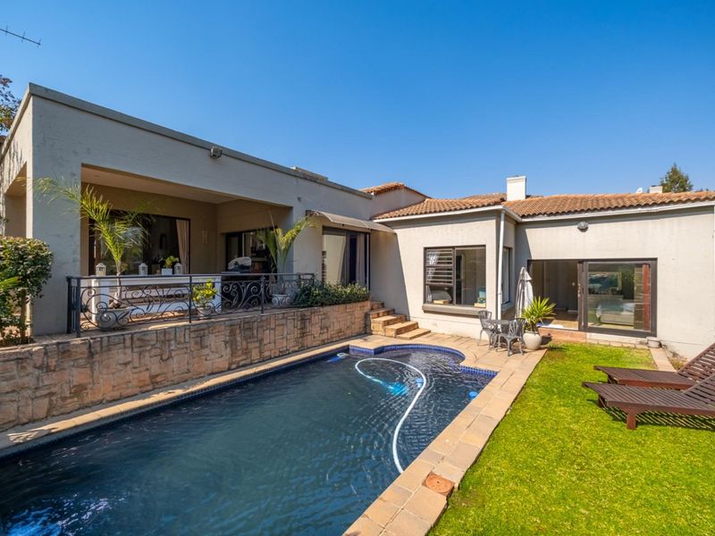 Rear exterior view featuring a swimming pool, stone retaining wall, covered patio area, and well-maintained garden space.