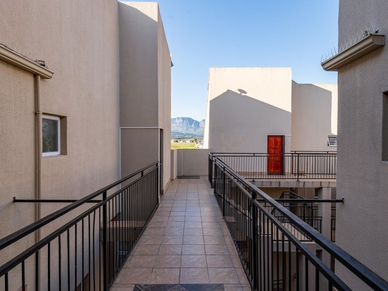 An exterior elevated walkway with tiled flooring and metal balustrades connecting apartment building sections, featuring views of the surrounding landscape.