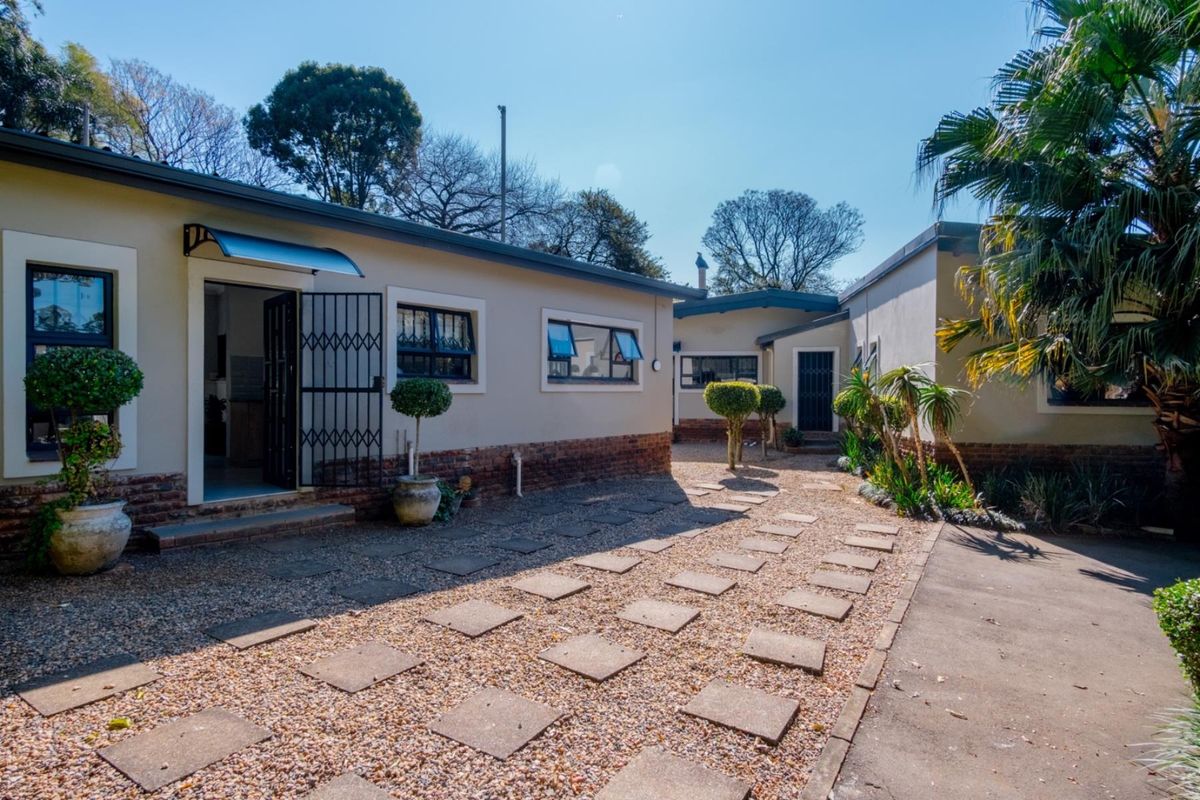 A well-maintained rear exterior view featuring a brick and plaster facade, paved walkway, and gravel garden area with security gates and window awnings.
