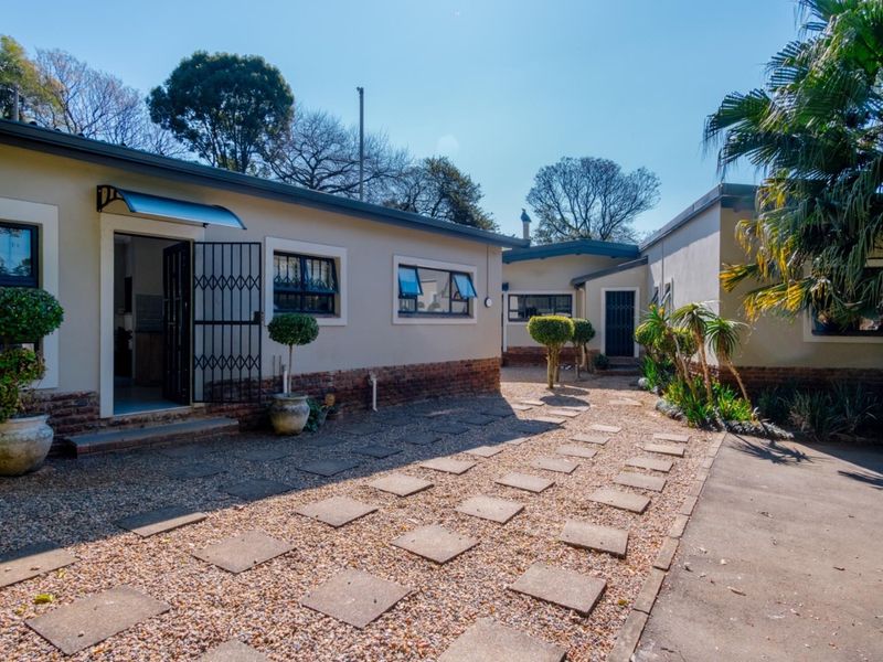 A well-maintained rear exterior view featuring a brick and plaster facade, paved walkway, and gravel garden area with security gates and window awnings.