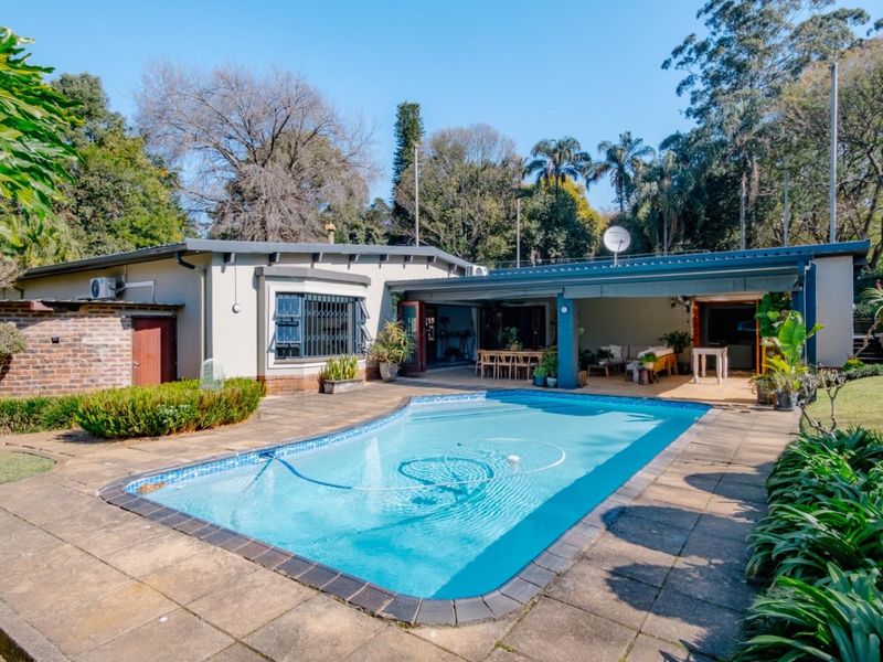 A well-maintained outdoor pool area featuring a paved deck, a covered patio space, and established garden surroundings.
