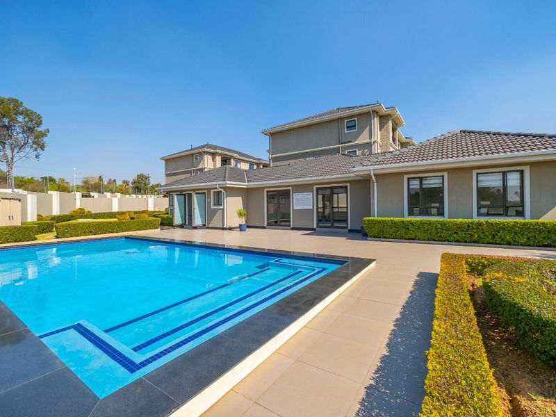 A well-maintained communal swimming pool area featuring a tiled deck, manicured hedges, and modern apartment building architecture with a tiled roof.