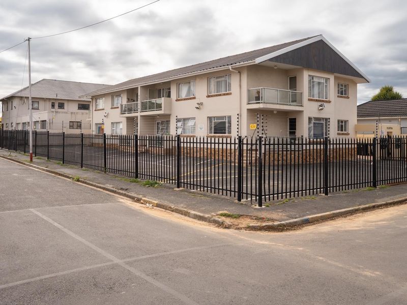 Exterior view of a multi-story apartment complex featuring a paved parking area, secure perimeter fencing with electric wires, and private balconies.