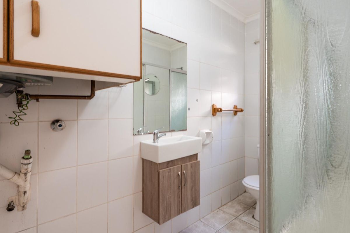 A compact bathroom featuring a wall-mounted vanity, shower enclosure, and toilet, with visible plumbing and dated finishes requiring modernization.