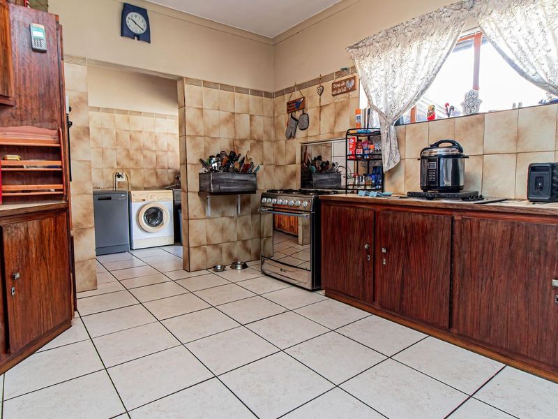 A functional kitchen featuring wooden cabinetry, tiled splashbacks, and tiled flooring, with an adjacent scullery area visible in the background.