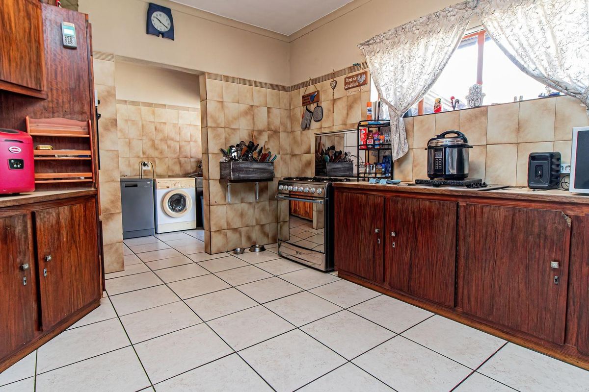 A functional kitchen featuring wooden cabinetry, tiled splashbacks, and tiled flooring, with an adjacent scullery area visible in the background.