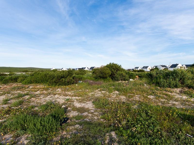 A view of a vacant residential plot featuring natural coastal vegetation and sandy terrain with surrounding neighborhood homes in the distance.