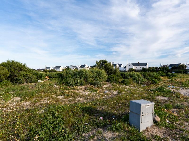 A vacant residential plot featuring natural vegetation and an existing electrical utility connection point, situated in a developed neighborhood.