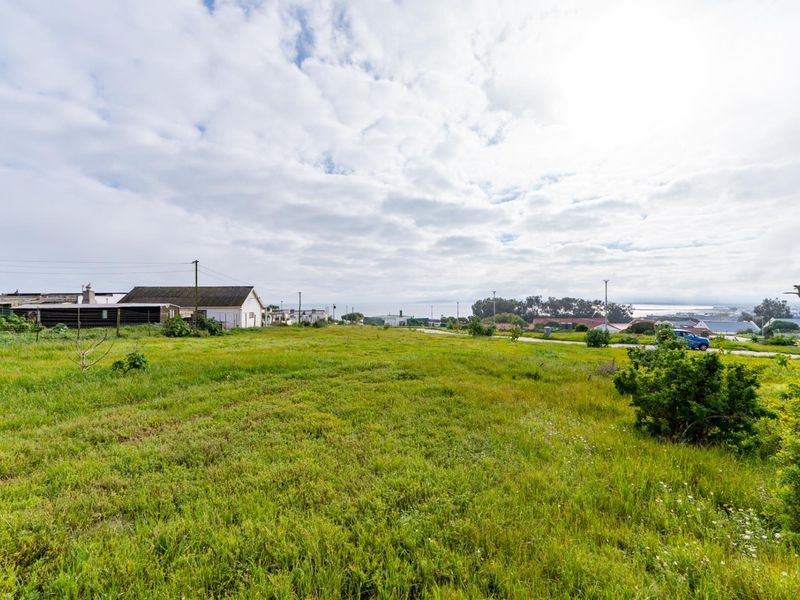 A wide-angle view of a vacant residential plot featuring open grassland with surrounding neighborhood infrastructure and distant coastal views.