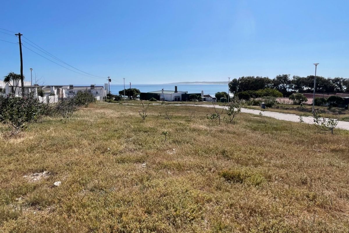 A view of a vacant residential plot featuring natural vegetation, utility poles, and a paved road with a distant ocean horizon.