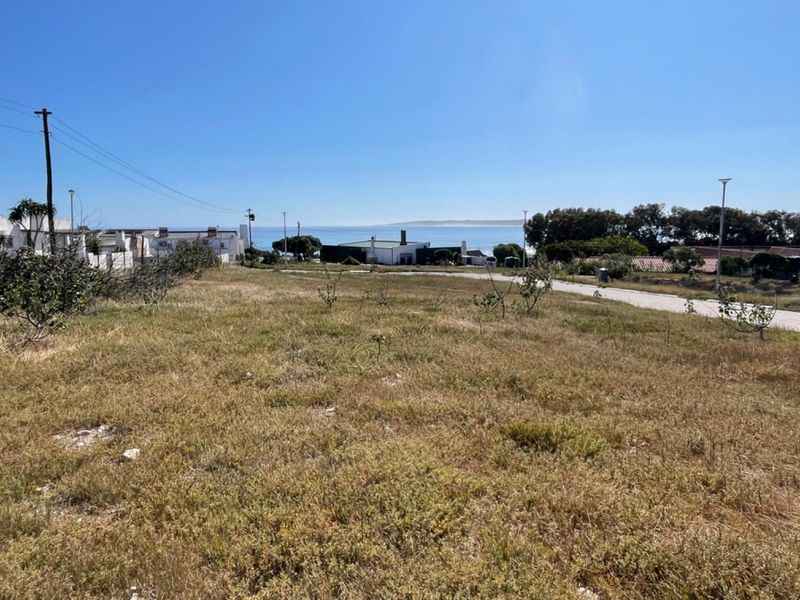A view of a vacant residential plot featuring natural vegetation, utility poles, and a paved road with a distant ocean horizon.
