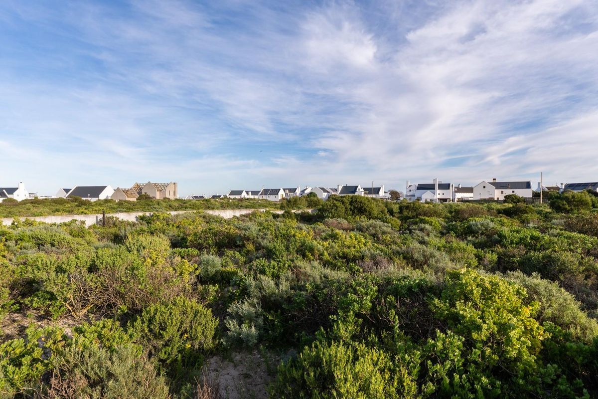 A wide-angle view of a vacant residential plot featuring native coastal vegetation and a boundary wall, with neighboring houses visible in the background.