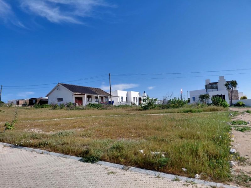 A vacant residential plot featuring natural grass cover, adjacent to a paved street with established neighboring properties in the background.