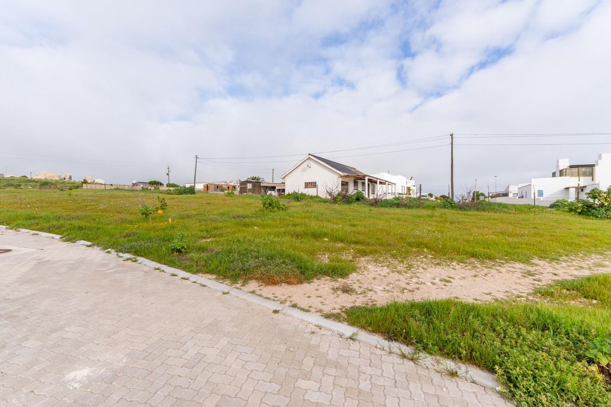 A vacant residential plot featuring a paved driveway, kerbing, and utility connections, with an existing structure visible in the background.