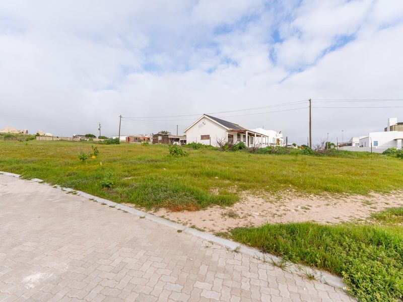 A vacant residential plot featuring a paved driveway, kerbing, and utility connections, with an existing structure visible in the background.