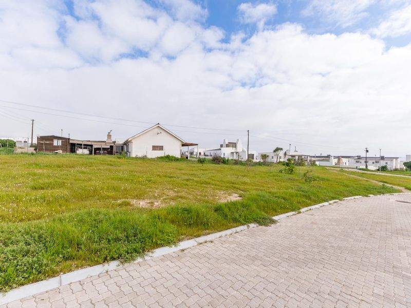 A vacant residential plot of land featuring a grassed surface, adjacent to a paved street with established neighborhood infrastructure in the background.