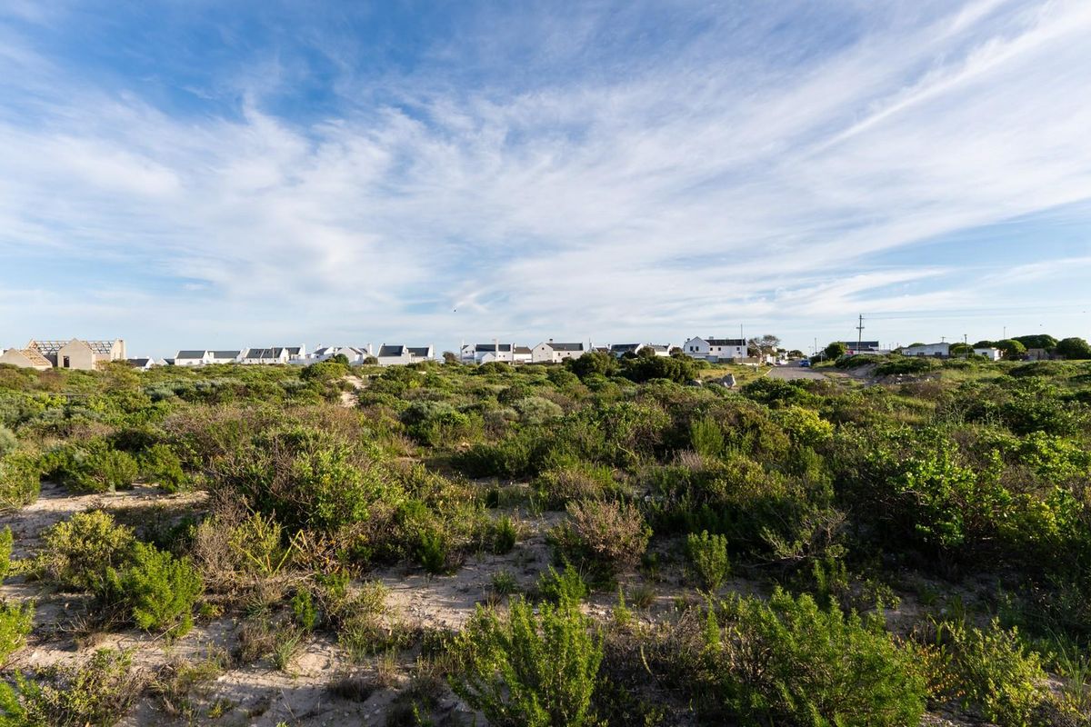 A wide-angle view of a vacant residential plot featuring natural coastal vegetation and sandy terrain with surrounding neighborhood homes in the distance.