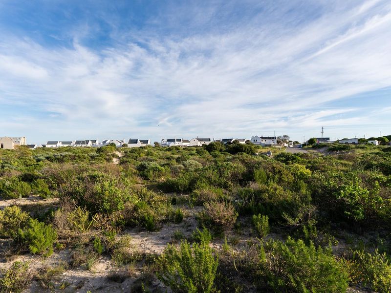 A wide-angle view of a vacant residential plot featuring natural coastal vegetation and sandy terrain with surrounding neighborhood homes in the distance.