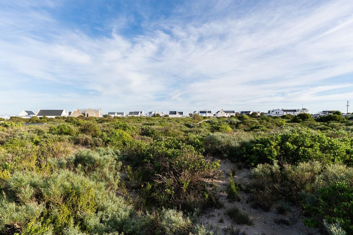 A wide-angle view of a vacant residential plot featuring natural coastal vegetation and sandy ground, with established homes visible in the background.