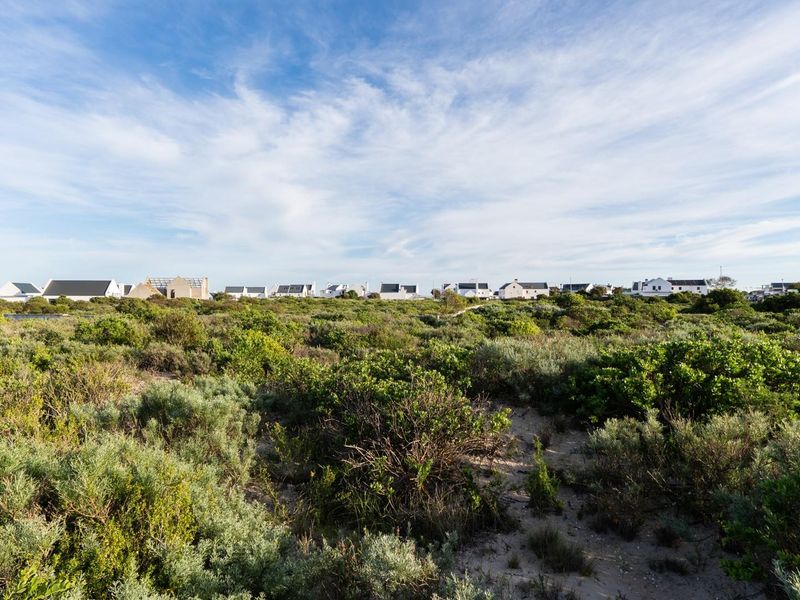 A wide-angle view of a vacant residential plot featuring natural coastal vegetation and sandy ground, with established homes visible in the background.