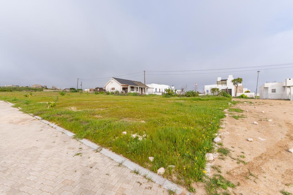 A vacant residential plot featuring grassed land, adjacent to a paved driveway and utility infrastructure in a quiet neighborhood setting.