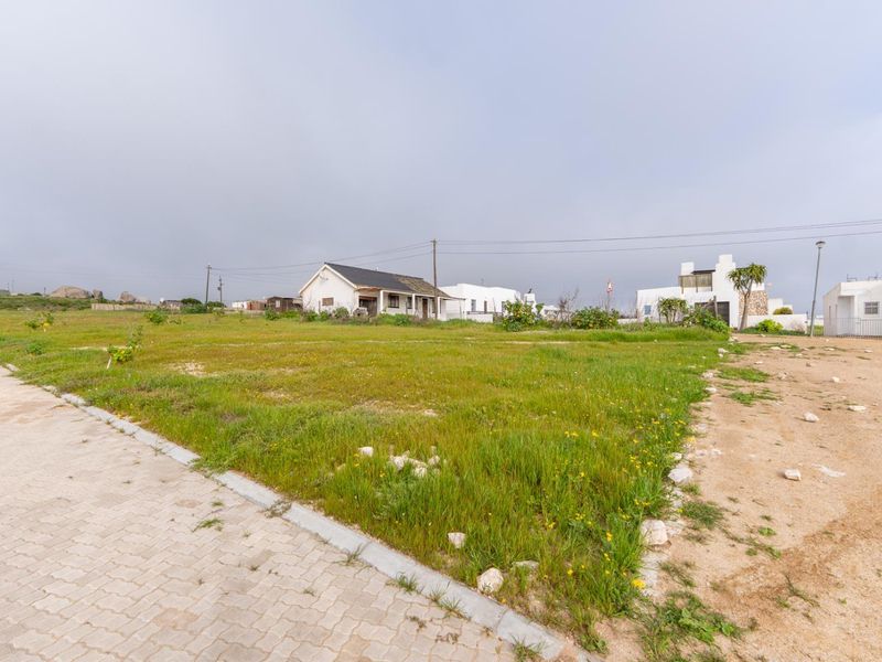 A vacant residential plot featuring grassed land, adjacent to a paved driveway and utility infrastructure in a quiet neighborhood setting.