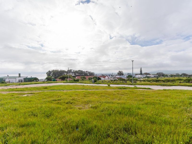 A vacant residential plot featuring grassy terrain with a view of the surrounding neighborhood and distant coastal landscape.