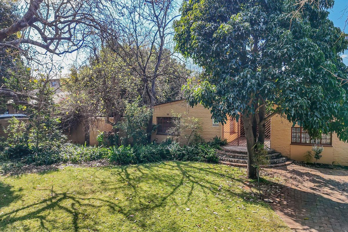 Rear view of the property featuring a brick facade, paved walkway, and established garden area with mature trees.
