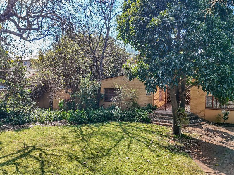 Rear view of the property featuring a brick facade, paved walkway, and established garden area with mature trees.