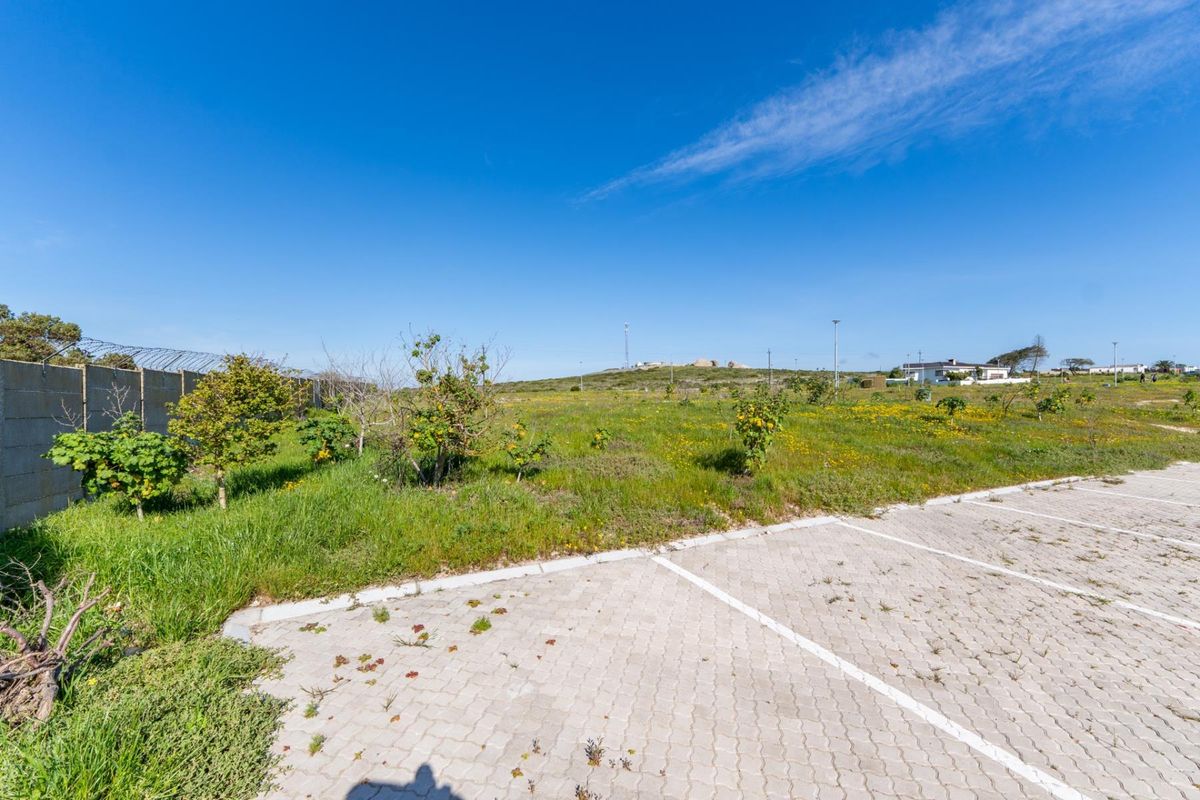 A vacant residential plot featuring a paved parking area, perimeter wall with security fencing, and natural vegetation.