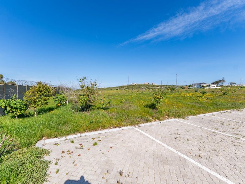 A vacant residential plot featuring a paved parking area, perimeter wall with security fencing, and natural vegetation.